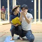 393335 Victoria Chen and Dai Tabuchi in rehearsals for My Neighbour Totoro © Manuel Harlan Victoria Chen wearing a hat and yellow bag sitting on Dai Tabuchi’s knee as he holds up something in his hands.